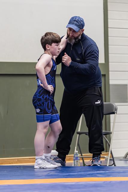 Een bebaarde coach in marine pet buigt zich voorover om een jonge worstelaar in een blauw tenue gerichte instructies te geven.