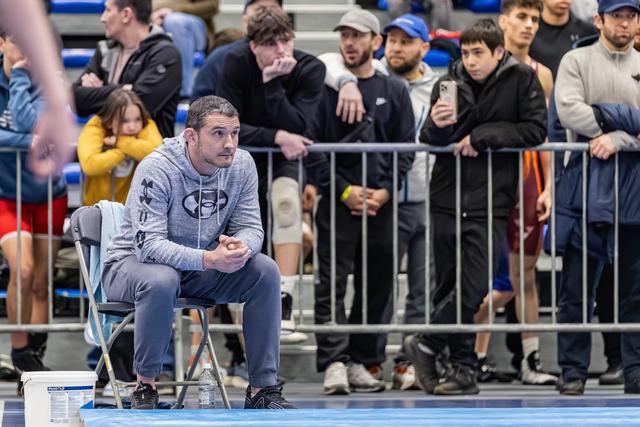 Coach in Under Armour hoodie zit gespannen ringside, handen gevouwen, met geconcentreerde blik op de wedstrijd gericht.