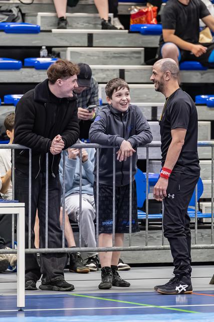 Kale coach en jonge tiener lachen samen met een oudere jongere naast een hek bij een indoorsportevenement.