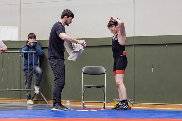 Coach geeft een handdoek aan een jonge worstelaar in een zwart tricot, die zijn arm strekt en uitrust tussen de gevechten door.