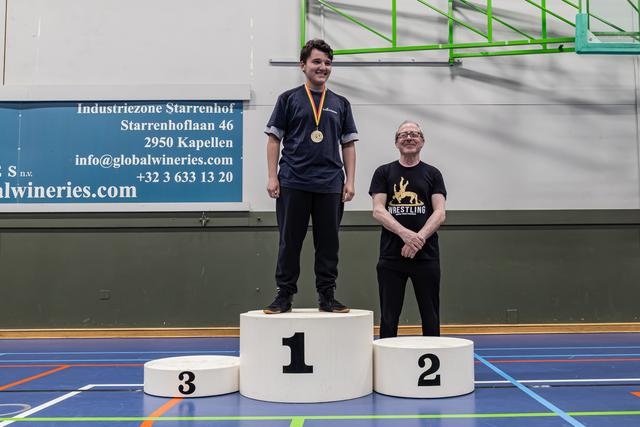Jonge man staat trots glimlachend op het podium met gouden medaille, oudere man staat naast het tweede podium in een worsteltshirt.