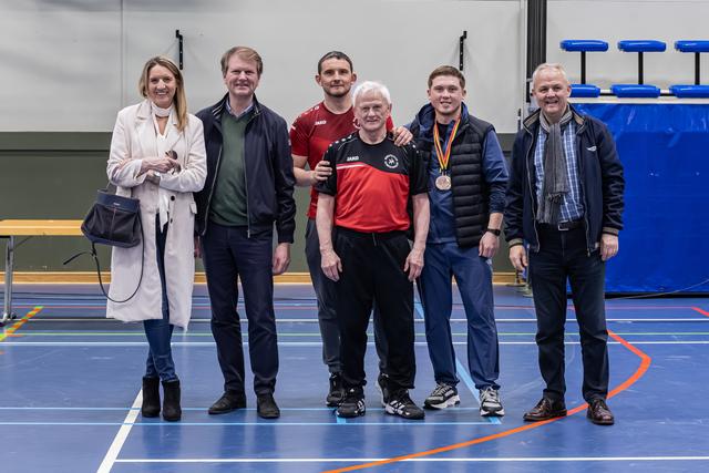 Zes mensen poseren trots in een sporthal; een jongeman draagt een bronzen medaille om zijn nek.