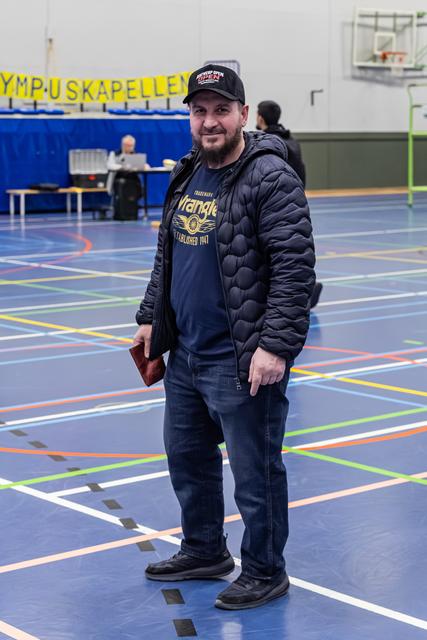 Bebaarde man in Wrangler-shirt en zwart jack staat ontspannen op een sporthalvloer en lacht naar de camera.