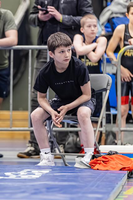 Jonge jongen in zwart shirt zit gespannen op een klapstoeltje langs de mat, handen gevouwen, en kijkt geconcentreerd toe.