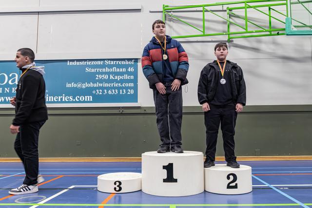 Twee jongens staan op plaatsen 1 en 2 van het podium met medailles, terwijl een derde langs positie 3 loopt in een sporthal.