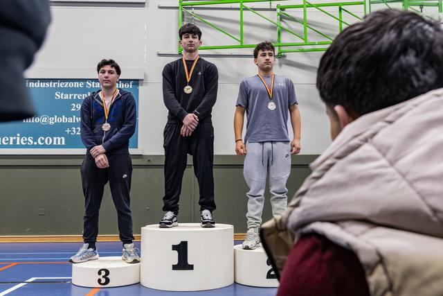 Drie jonge mannelijke atleten staan op het podium op plaatsen 1, 2 en 3, met medailles om, met een ernstige blik in een sporthal.