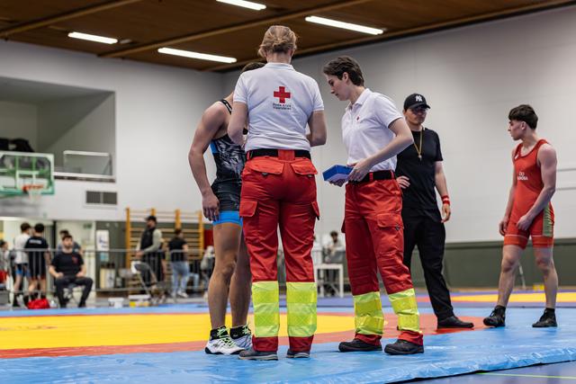 Twee Rode Kruis-hulpverleners overleggen met een worstelaar op de mat tijdens een pauze, terwijl een scheidsrechter en atleet toekijken.