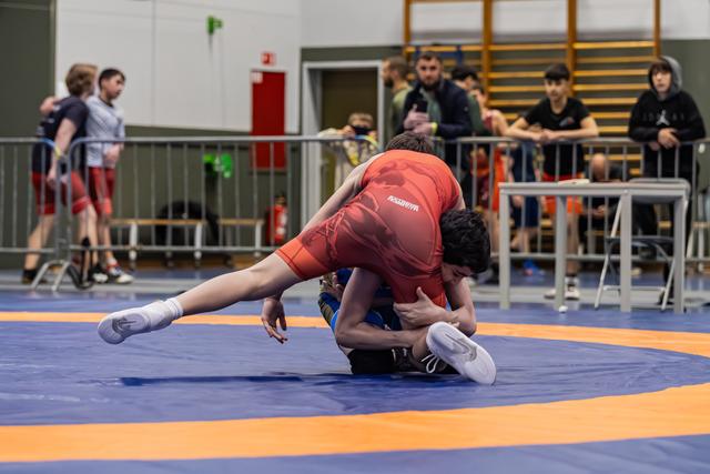 Worstelaar in rood pak voert een takedown uit en drukt tegenstander op de blauwe mat terwijl toeschouwers toekijken.