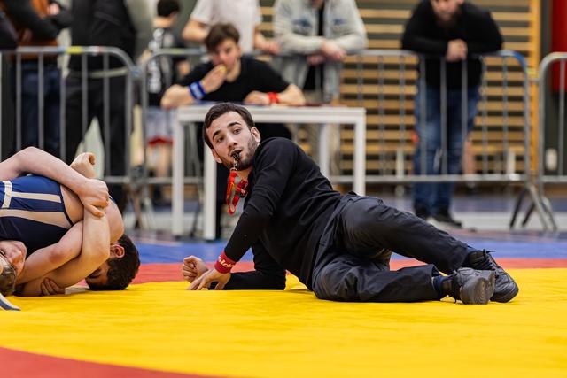Scheidsrechter ligt op gele mat en fluit terwijl hij twee worstelaars op de voet volgt tijdens een indoortoernooi.