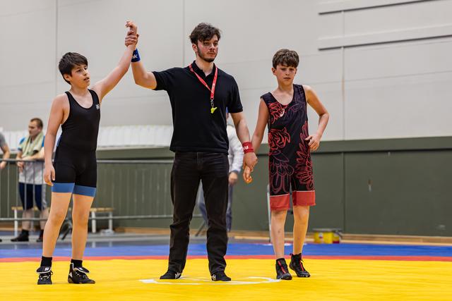 Scheidsrechter heft de hand van de jonge worstelaar omhoog als winnaar, terwijl de verliezer teleurgesteld naast hem staat.