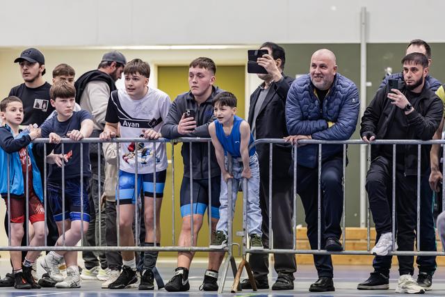 Coaches, atleten en ouders leunen op metalen hekken en kijken gespannen toe bij een indoorworsteltoernooi.