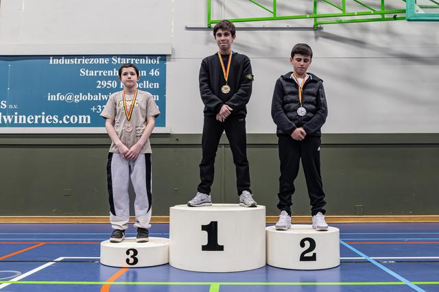 Drie jonge jongens staan op een podium met medailles in een sporthal. De winnaar kijkt ingehouden.