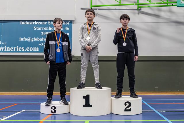 Drie jongens staan op het podium met goud, zilver en brons, allen gedecoreerd met een medaille in een sporthal.