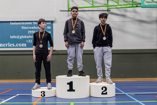 Drie jongens staan op een podium met medailles; de eerste staat het hoogst, de derde kijkt teleurgesteld.