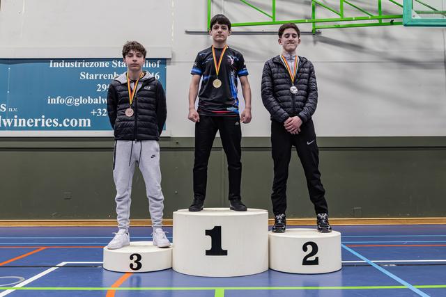 Drie jonge mannelijke worstelaars staan op een podium met brons, goud en zilver, met ernstige gezichten in een sporthal.