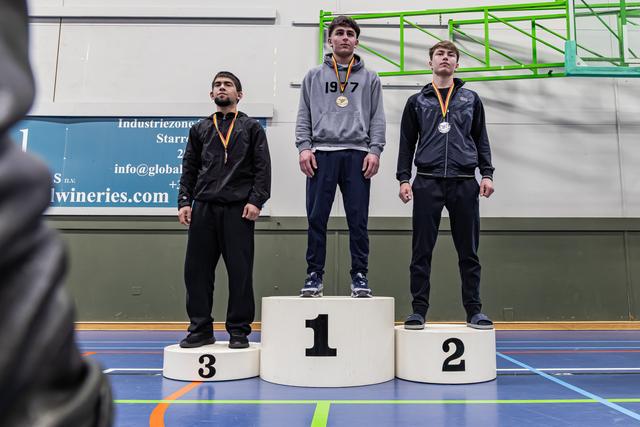 Drie jonge worstelaars staan op een podium met medailles. De gouden medaillewinnaar staat in het midden en kijkt vastberaden.