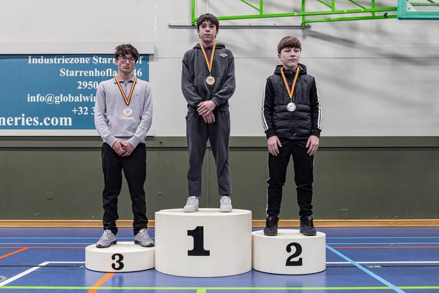 Drie jonge mannelijke atleten staan op een podium, eerste plaats in het midden met gouden medaille, tweede en derde met zilver en brons, in een sporthal.