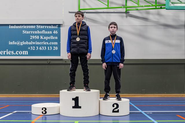 Twee jonge jongens staan op een podium op de eerste en tweede plaats met medailles in een Belgische sporthal.