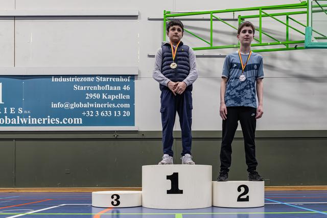 Twee jonge jongens staan op een podium, de eerste met gouden medaille, de tweede met zilver, in een sporthal.