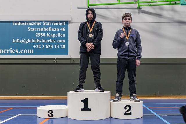 Twee jonge jongens staan op het podium; de gouden medaillewinnaar staat rustig terwijl de zilveren medaillewinnaar trots zijn medaille toont.