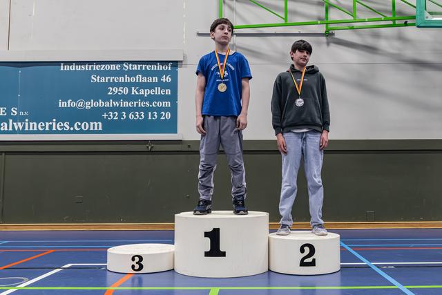 Twee jongens staan op een podium op de eerste en tweede plaats met medailles, met een ernstige blik in een sporthal.