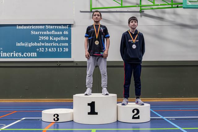 Twee jonge jongens staan op het podium als goud- en zilvermedaillewinnaar, met een ernstige blik in een sporthal.