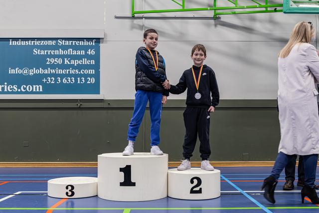 Twee jongens schudden elkaar de hand op een ereplaats, beiden lachend met medailles om hun nek in een sporthal.