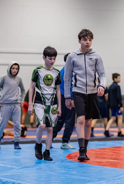 Twee jonge worstelaars lopen over de mat; de kleinste in groen tenue kijkt neerslachtig, de langste in een Nike hoodie loopt naast hem.