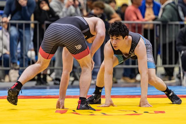 Twee jonge worstelaars hurken gespannen tegenover elkaar op een gele mat, de voorste atleet toont scherpe concentratie.