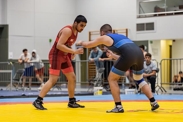 Twee worstelaars in rode en blauwe pakken staan tegenover elkaar op een gele mat in een sporthal, toeschouwers op de achtergrond.