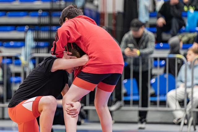 Twee jonge worstelaars in een strakke greep op de mat, beiden naar voren gebogen met intense concentratie tijdens een indoorwedstrijd.