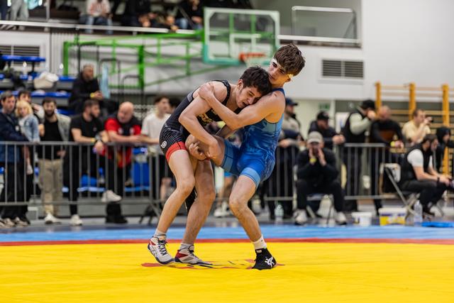 Twee jonge worstelaars verwikkeld in een intense greep op een gele mat, toeschouwers kijken toe in een volle sporthal.