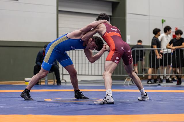 Twee jonge worstelaars gaan vol in de clinch op de mat, de atleet in blauw vecht verbeten tegen zijn Belgische tegenstander in donkerrood.