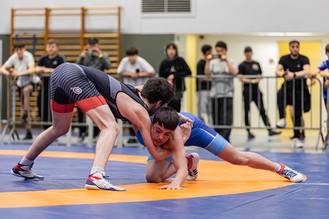 Jonge worstelaar in blauw tenue probeert een takedown te ontwijken op de oranje mat, toeschouwers kijken toe achter de hekken.