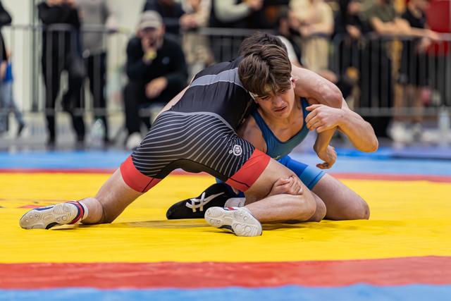 Jonge worstelaar in blauwe singlet trekt gefocust gezicht terwijl hij tegenstander in zwart gestreepte singlet beheerst op gele mat.