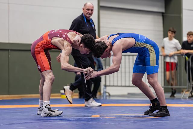 Twee jonge worstelaars grijpen elkaars handen in een gespannen duel op de blauwe mat, terwijl de scheidsrechter toekijkt.