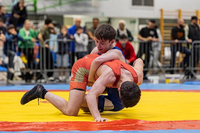 Jonge worstelaar in blauw houdt tegenstander in rood op de gele mat tijdens een indoortoernooi.