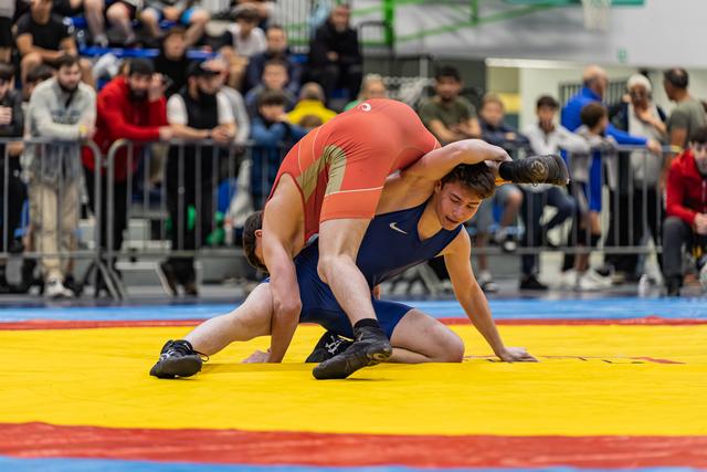 Worstelaar in blauw tenue verzet zich tegen tegenstander in rood die een takedown probeert op de gele mat.