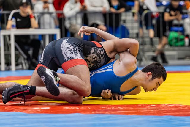 Worstelaar in blauwe singlet wordt door tegenstander in zwarte tijgerprint singlet met gezicht naar beneden op de mat gedrukt.