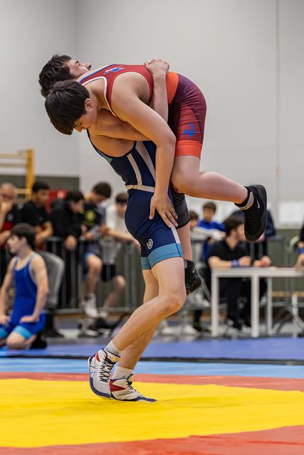 Jonge worstelaar in marineblauw tenue tilt zijn tegenstander volledig van de mat tijdens een indoortoernooi.