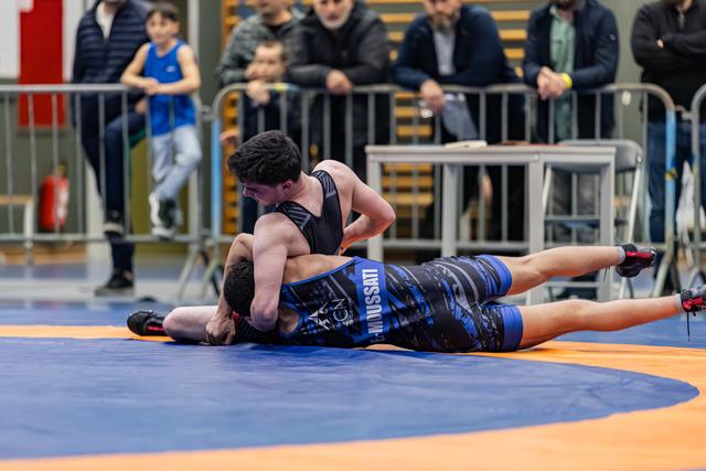 Atleet in zwart tenue zet tegenstander in blauw 'Mussati' tenue vast op de mat, toeschouwers kijken toe achter hekken.