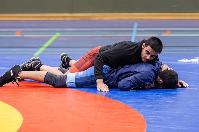 Jonge worstelaar in zwart shirt houdt tegenstander in blauw wedstrijdpak op de mat, stevig in controlepositie.