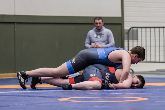 Jonge worstelaar in blauw tenue houdt tegenstander op de mat terwijl een coach aandachtig toekijkt op de achtergrond.