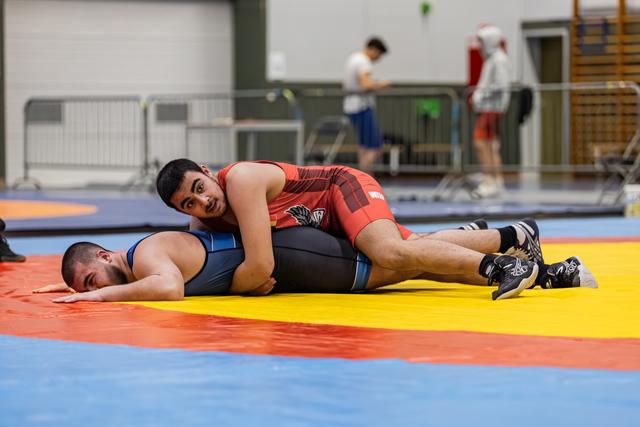 Jonge worstelaar in rood tenue houdt tegenstander op de mat, met geconcentreerde blik in een sporthal.