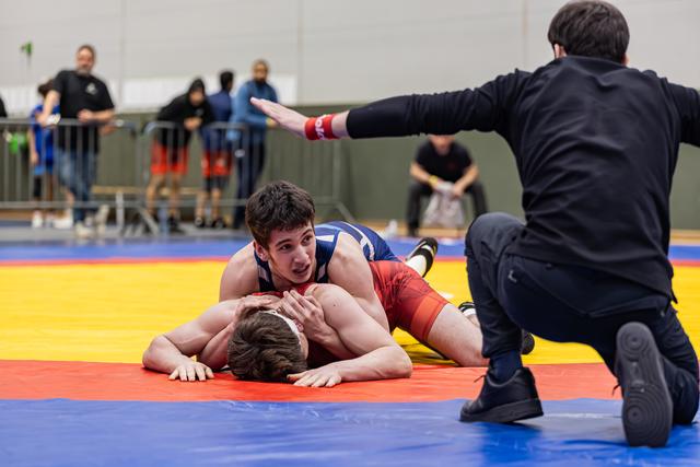 Jonge worstelaar in blauw drukt tegenstander plat op de mat terwijl de scheidsrechter knielt en zijn hand opheft.