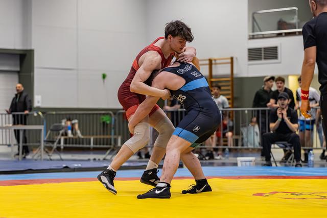 Twee worstelaars in een intense greep op een gele mat, atleet in rood shirt dringt agressief vooruit in een overdekte sporthal.