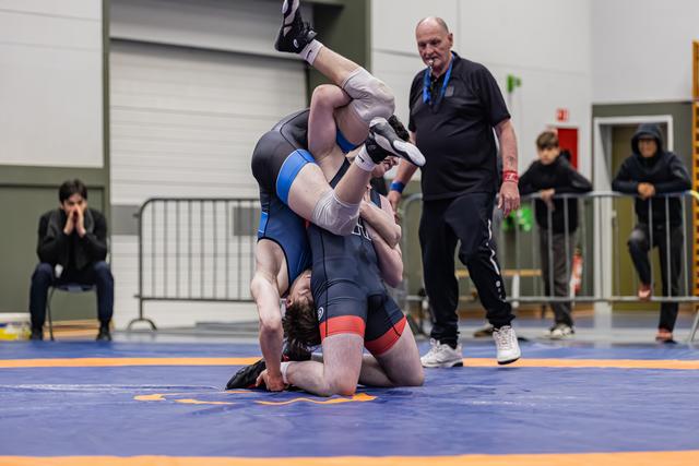 Twee worstelaars verwikkeld in een greep op de mat terwijl een kale scheidsrechter glimlachend toekijkt in een sporthal.