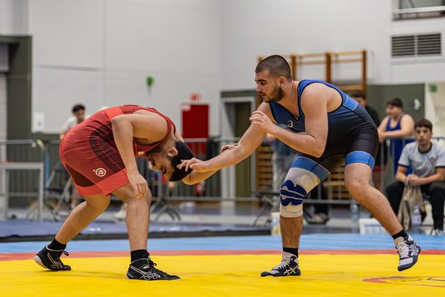 Twee worstelaars in actie op een gele mat; de atleet in blauw dringt op, terwijl zijn tegenstander in rood diep vooroverbuigt.