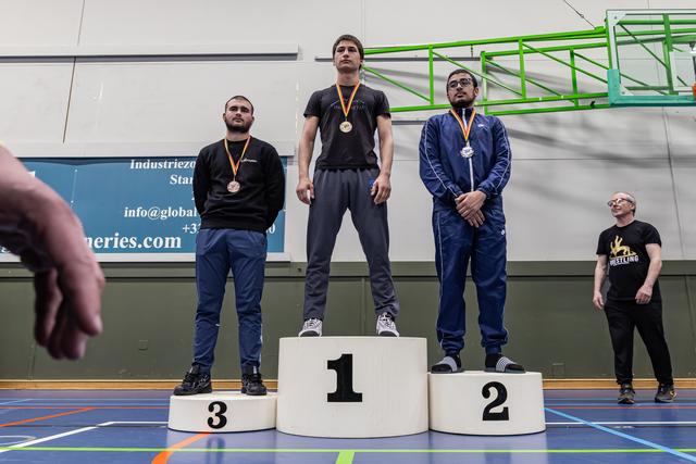 Drie mannelijke worstelaars staan op het podium, goudmedaillewinnaar in het midden op plaats 1, geflankeerd door zilver en brons in een sporthal.