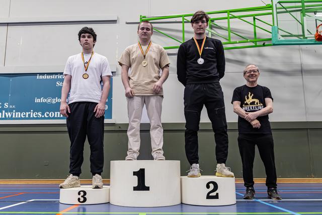 Drie jonge worstelaars staan op het podium (1e, 2e, 3e) met medailles om, terwijl een lachende coach naast hen staat in een sporthal.